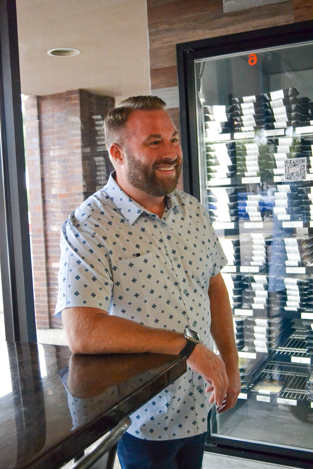 Smiling man leaning on a counter by a window in a patterned shirt.